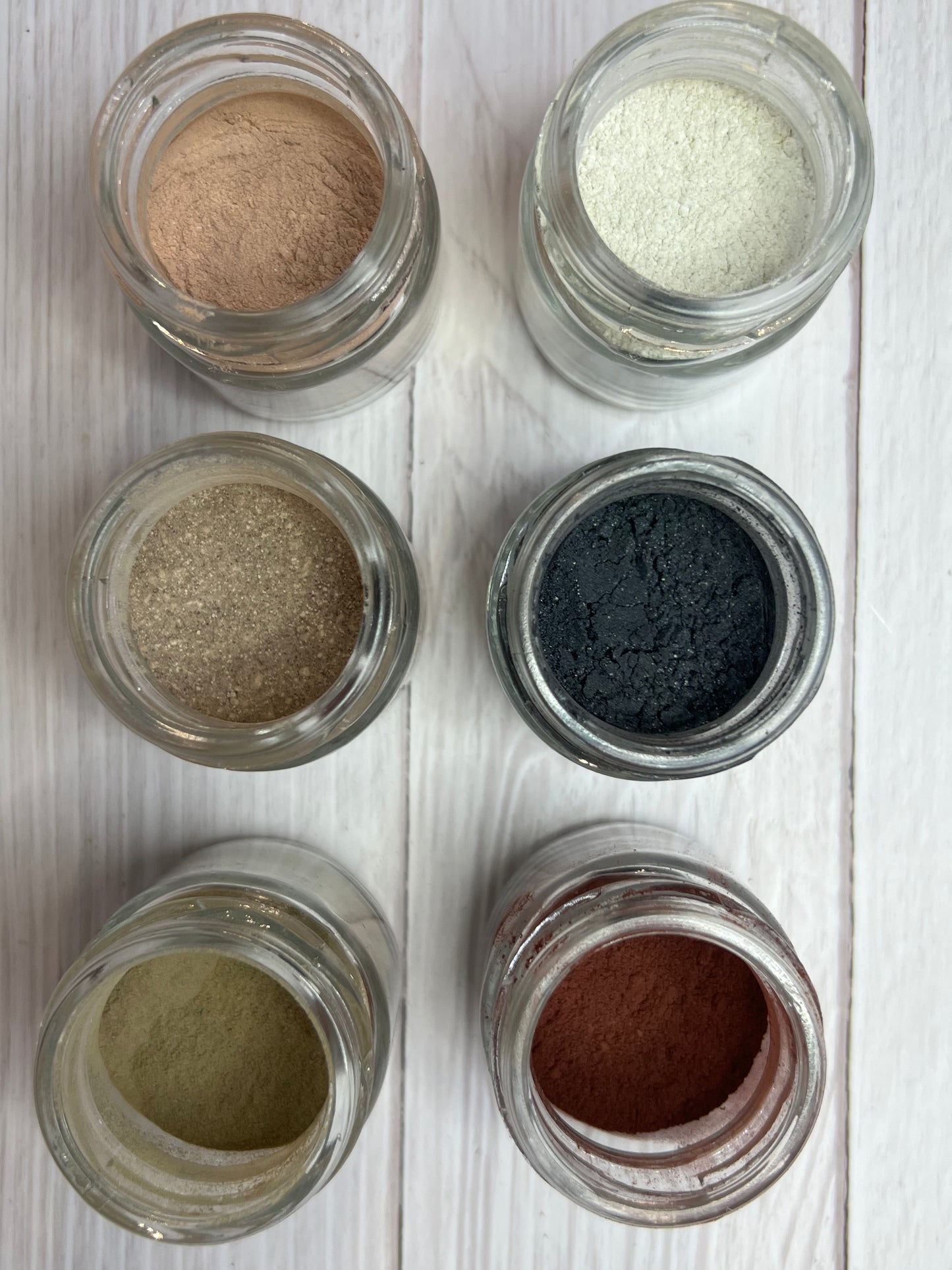 Six small glass bowls filled with different natural powders in earthy shades (green, yellow, black, brown, red, beige) arranged on a grey background.