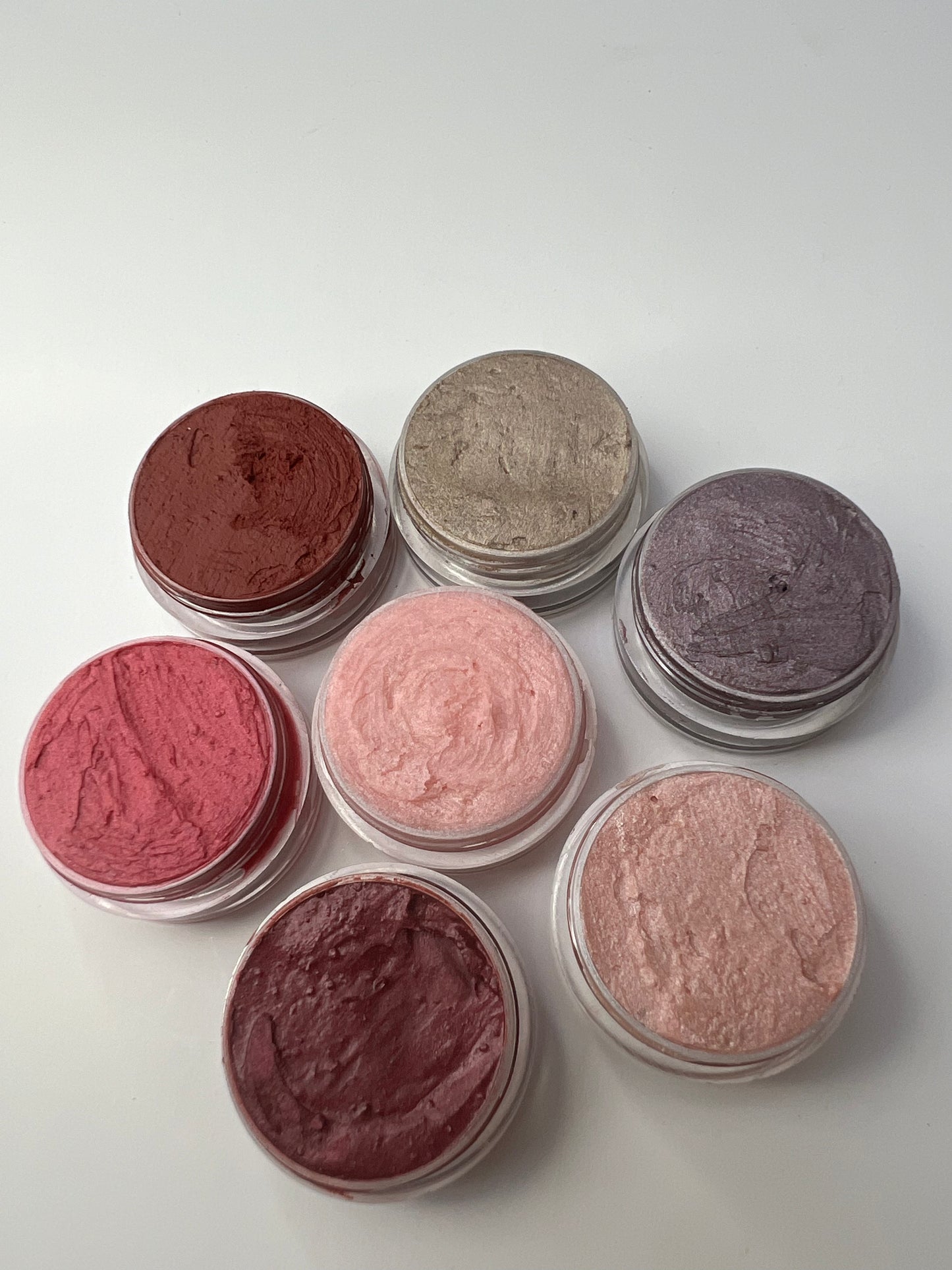 Seven handmade lip balms in different colors and shimmer finishes, packaged in small round tins, displayed on a white background.