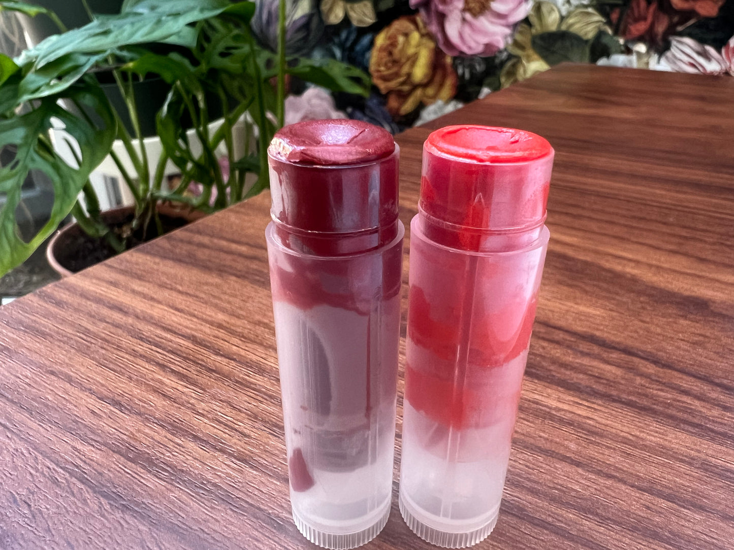 Two clear lip balms with red caps on a wooden surface with plants in the background