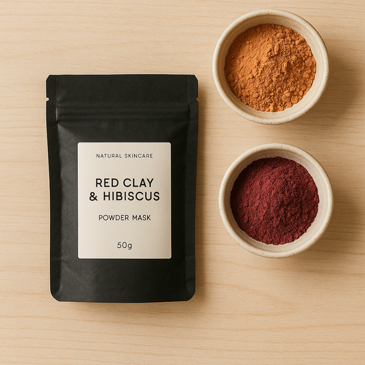 A black resealable pouch labeled “Red Clay & Hibiscus Powder Mask, 50g” placed on a wooden surface, with two small bowls of red and orange powders beside it.