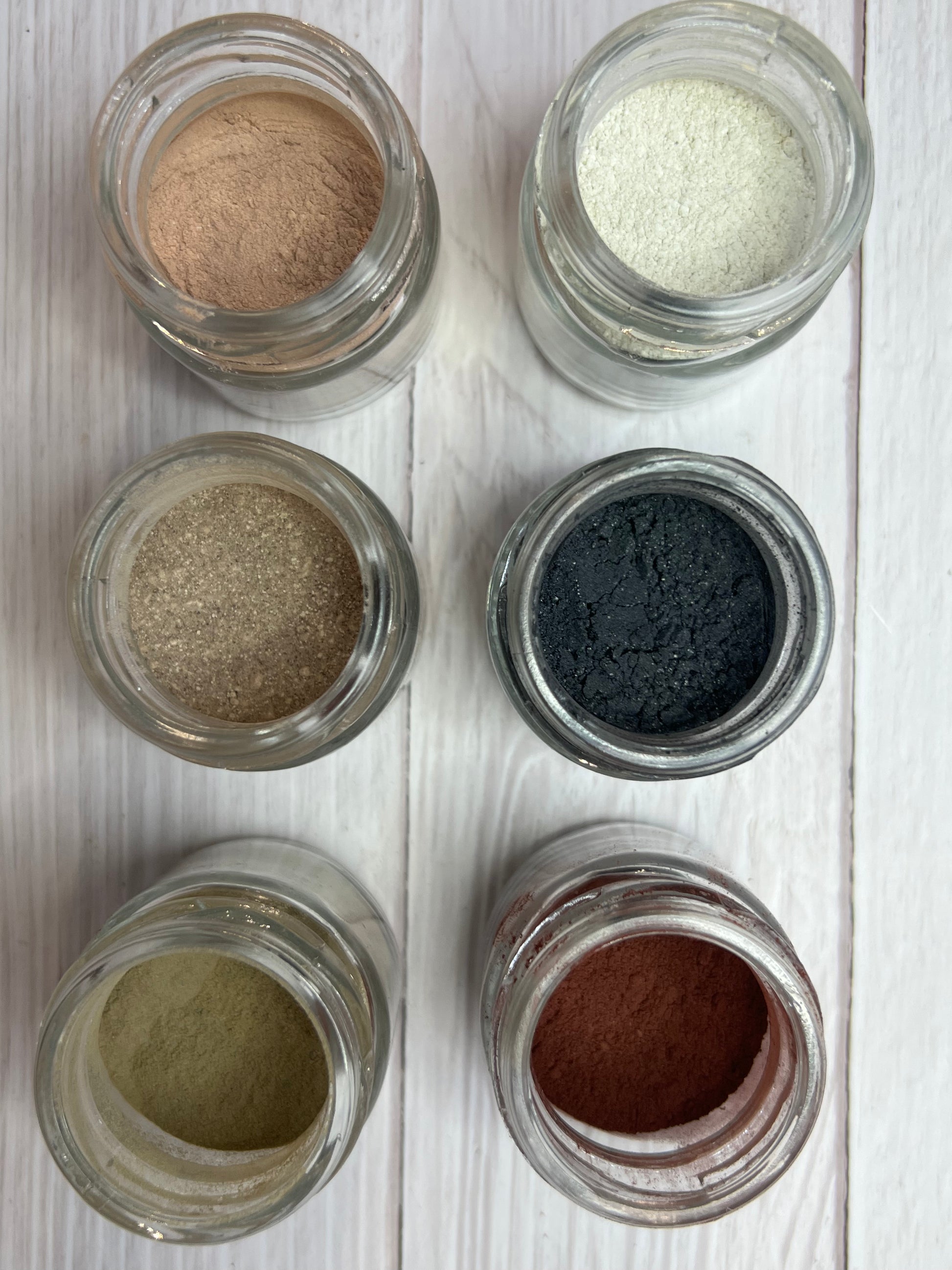 Six small glass bowls filled with different natural powders in earthy shades (green, yellow, black, brown, red, beige) arranged on a grey background.