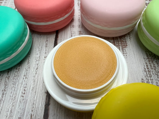 Assorted pastel macaron-shaped lip balm pots arranged on a rustic wooden board.