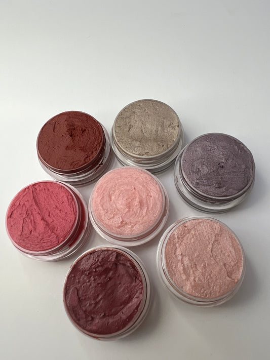 Seven handmade lip balms in different colors and shimmer finishes, packaged in small round tins, displayed on a white background.