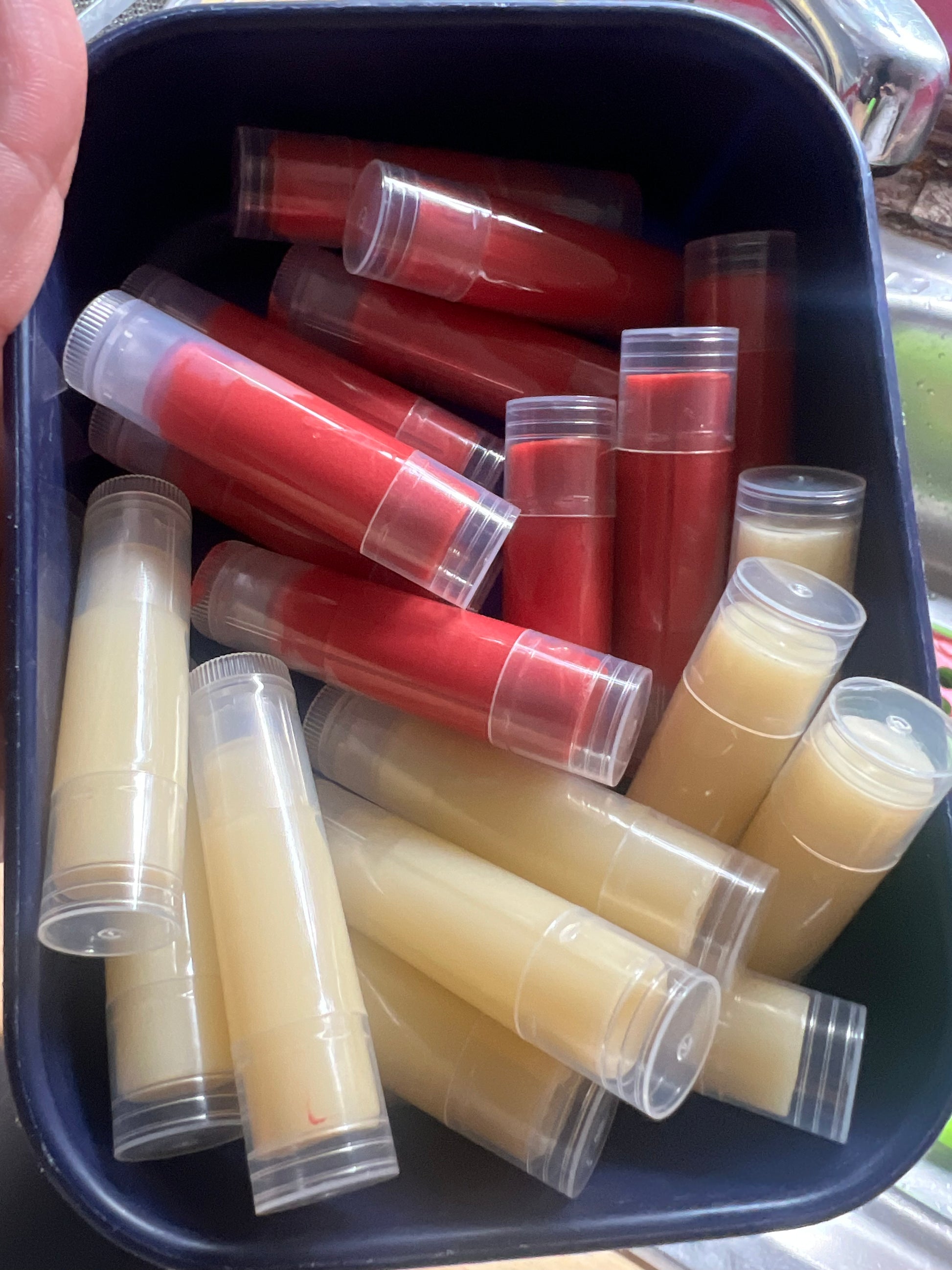 Close-up of a dark container filled with a mix of unlabeled lip balm tubes, some with a natural creamy yellow color and others tinted red. 