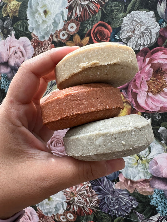 Hand holding three stacked shampoo bars in different colors against a floral background.
