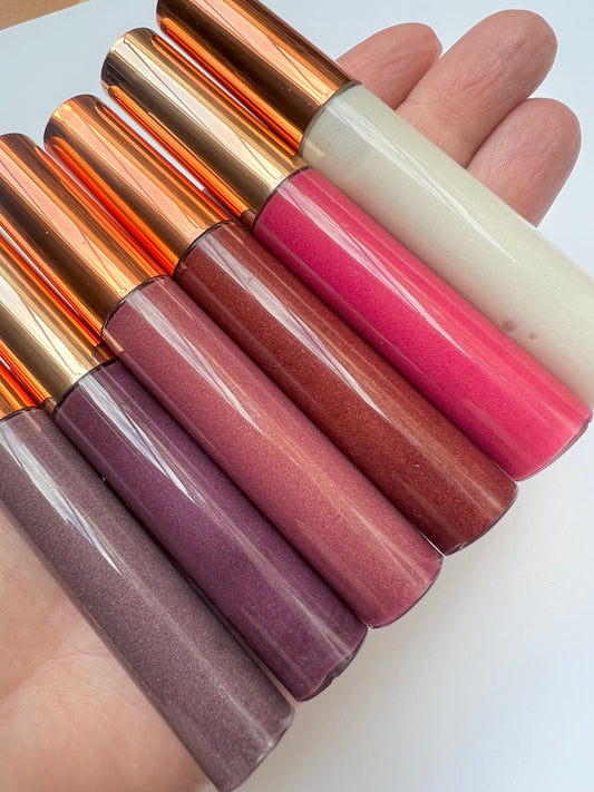 Hand holding six tubes of handmade lip gloss in various shades, including purple, pink, nude, and clear, with rose-gold caps.