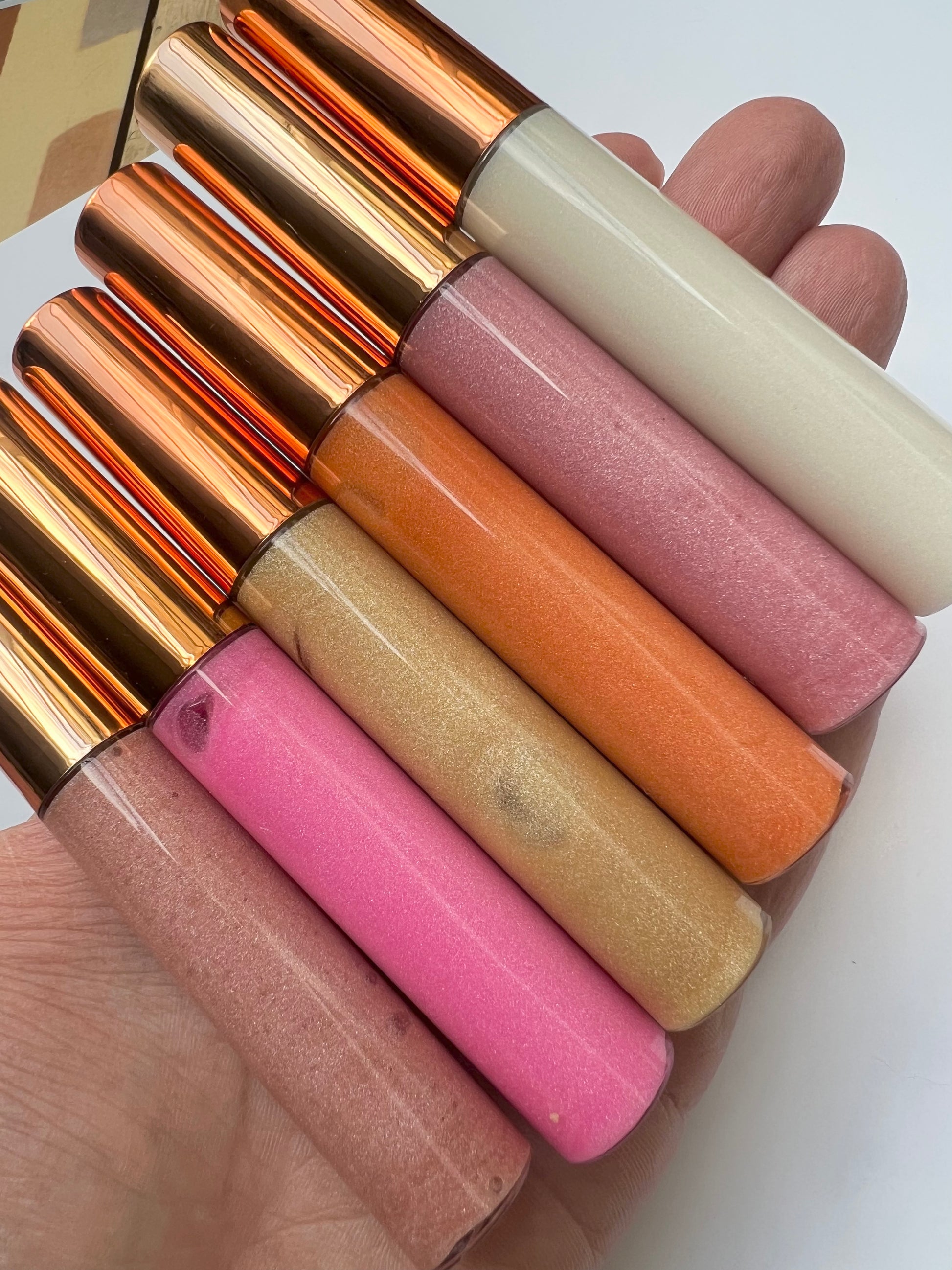 Five handmade lip gloss tubes in shades of pink, nude, and bronze with rose-gold caps, displayed in a hand.