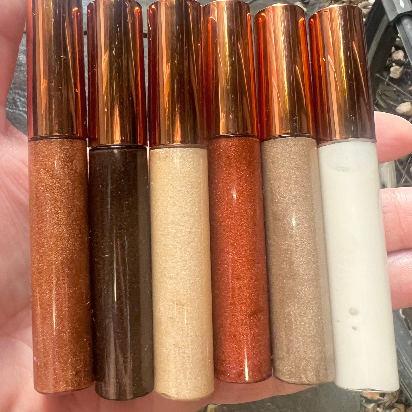 Row of six handmade lip gloss tubes in natural shades from nude to bronze, with rose-gold caps.