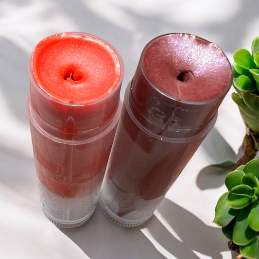 Two tinted lip balms standing upright — one in deep berry shade and one in coral red, with a green succulent in the background.